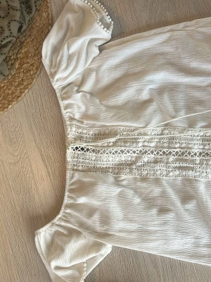 Vit offshoulder blus med spetsdetaljer - Söt vit offshoulder blus med dekorativa spetsdetaljer framtill och små bollar längs ärmslut. Blusen har en luftig och avslappnad passform, perfekt för sommardagar. Materialet känns lätt och mjukt mot huden.