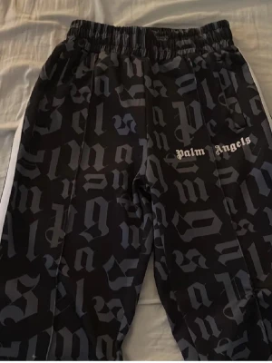 Palm Angels svarta mjukisbyxor - Säljer ett par svarta mjukisbyxor från Palm Angels med grått gotiskt mönster och vit logga på ena benet. Byxorna har resår i midjan och vita ränder längs sidorna. Perfekta för en streetwear-look. Varför jag säljer så billigt är eftersom palm angels loggan är lite dålig och det finns några få lösa trådar.
