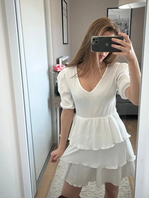 Vit volangklänning med v-ringning - Söt vit klänning med v-ringad hals och puffärmar. Klänningen har flera lager volanger som ger en luftig och romantisk känsla. Tillverkad i ett lätt tyg, perfekt för varma dagar. Passformen är normal och längden är kort.