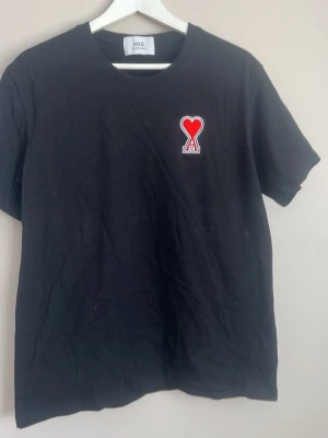 Svart t-shirt från Ami med hjärtdetalj - Svart t-shirt från Ami med klassisk passform och rund hals. På bröstet sitter en broderad röd hjärtdetalj med bokstaven A. Tillverkad i mjuk bomull som känns skön mot huden. Perfekt för en stilren och trendig look.