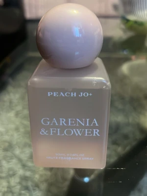 Peach Jo+ Gardenia & Flower parfym - En stilren parfymflaska i ljusrosa med rund kork och rektangulär form. Märkt med Peach Jo+ och doften Gardenia & Flower. Flaskan rymmer 90 ml och är tillverkad i glas med en modern, minimalistisk design.