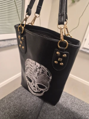 Svart handväska med döskallemotiv och guld detaljer  - Cool svart handväska i konstläder med ett stort vitt döskallemotiv framtill. Väskan har guldfärgade metalldetaljer, nitar och dubbla axelband. Perfekt för dig som gillar edgy och unik stil.