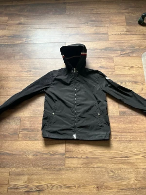 Svart moncler vindjacka  - Snyggt svart vindjacka med huva och dragkedja framtill. Jackan har två fickor med knappar och en cool detalj med röd och vit rand på huvan. Perfekt för blåsiga dagar och enkel att matcha med olika outfits.