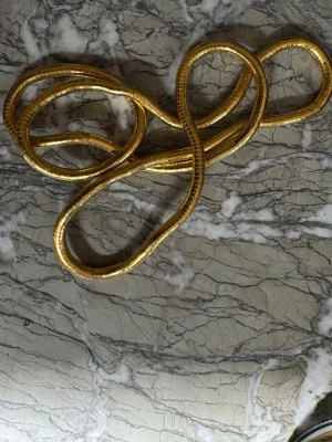 Guldigt statement-halsband/armaband - Unikt halsband i guldtonad metall med en tjock, ormliknande kedja som kan formas till olika knutar. Halsbandet har en glänsande finish och ger en riktigt cool och modern look. Perfekt för dig som vill sticka ut och addera lite edge till din outfit.