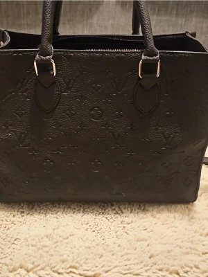 Svart handväska - Säljer en svart handväska  i präglat läder med klassiskt monogrammönster. Väskan har både handtag och axelrem, samt silverfärgade metalldetaljer. Perfekt för dig som vill ha en stilren och lyxig accessoar.