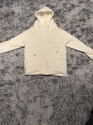 Vit cardigan  - Stilren vit pufferjacka med quiltad framsida och dragkedja. Jackan har två fickor med dragkedja och en praktisk huva. Perfekt nu till våren. Ordinarie pris ligger den på 1000kr 