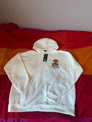 Vit hoodie med nalletryck från Crime - Säljer en vit hoodie från Crime med ett stort tryck av en söt nallebjörn i streetwear-stil på bröstet. Hoodien har huva och känguruficka framtill. Materialet känns mjukt och skönt, perfekt för chill dagar. Stora bokstäver 'CRIME' under trycket ger en cool vibe.