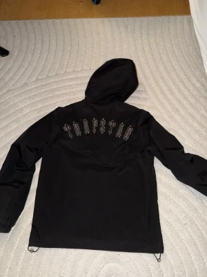 Svart Trapstar vindjacka med huva - Snygg svart vindjacka från Trapstar med stor logga i vitt på ryggen och rund patch på bröstet. Jackan har dragkedja framtill, två sidofickor med knappar och justerbar huva. Perfekt för streetwear-stil och enkel att matcha.