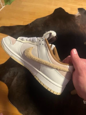 Nike Dunk Low  - Unika Nike Dunk Low  sneakers i beige och vitt med detaljer i mocka och läder. Swoosh-loggan är i lurvig beige textil och skon har bruna och vita inslag samt broderad blomma på hälen. Perfekt för dig som gillar limited edition och streetwear.