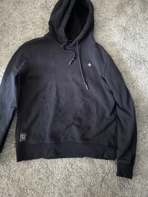 Mörkblå hoodie från Morris Stockholm  - Snygg mörkblå hoodie från Morris Stockholm i storlek XS. Klassisk modell med huva och dragsnören, liten logga broderad på bröstet och patch nertill. Tillverkad i mjukt och bekvämt material, perfekt för en avslappnad stil.