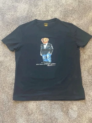 Svart Polo Bear t-shirt från Ralph Lauren - Svart t-shirt från Ralph Lauren med ikoniska Polo Bear-trycket på bröstet. Klassisk rund hals och korta ärmar. Tillverkad i mjuk bomull för skön känsla och snygg passform. Perfekt för dig som gillar streetwear och stilrena prints. Säljer pga fel storlek