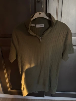 Olivgrön stickad tröja Selected Homme - Säljer en olivgrön stickad tröja från Selected Homme med korta ärmar och halvzip framtill. Tröjan har ribbad struktur och en hög krage som kan bäras öppen eller stängd. Perfekt för dig som gillar stilrena och moderna plagg.