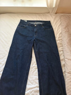 Mörkblåa pösiga jeans med hög midja - Säljer ett par mörkblå jeans med rak och pösig passform och hög midja. Klassisk femficksmodell med knapp och dragkedja framtill. Jeansen har breda ben och är tillverkade i ett kraftigt denimtyg, perfekta för en avslappnad och trendig look.