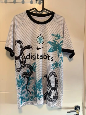 Inter Milan matchtröja med ormmönster - Fin Inter Milan matchtröja från Nike med svart ormmönster och turkosa blad på vit bakgrund. Tröjan har svarta detaljer vid hals och ärmslut, digitalbits-logga på bröstet och ett unikt geometriskt mönster på ryggen. Perfekt för fotboll eller streetwear.