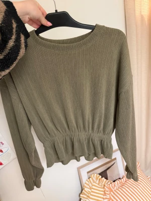 Olivgrön ribbad tröja med volang - Trendig olivgrön tröja med ribbad struktur och volangdetalj nedtill. Tröjan har rund halsringning, långa ärmar och smockad midja som ger en snygg siluett. Perfekt för dig som gillar en avslappnad men ändå söt stil. Har en liten fläck inne i tröjan, inget som syns utanför 