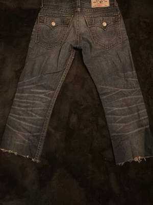 True Religion jeans  - Säljer ett par blåa jeans från True Religion med klassiska kontrastsömmar och stora bakfickor med lock och knappar. Jeansen har slitningar och råa, avklippta benslut för en edgy look. Materialet är denim i bomull och passformen är rak. 90 cm långa