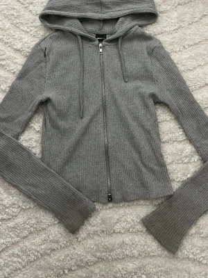 Grå ribbad hoodie från Gina Tricot - Säljer en grå ribbad hoodie från Gina Tricot med dragkedja framtill och långa ärmar. Hoodien har en klassisk huva med dragsko och är gjord i ett mjukt, stretchigt material som sitter snyggt. Perfekt för en chill och stilren look.