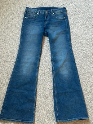 Blå bootcut jeans med fickor - Snygga blå bootcut jeans med klassiska fem fickor och knappgylf. Jeansen har en lätt utsvängd passform från knät och ner, vilket ger en retrovibe. Perfekta att styla med sneakers eller boots för en cool look. Byxorna passar perfekt för en S med!