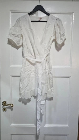 Vit omlottklänning med puffärm - Aldrig använd! Snygg vit omlottklänning med puffärmar och knytband i midjan. Klänningen har v-ringning och kort längd, perfekt för sommarens varma dagar. Tillverkad i ett lätt och luftigt tyg som ger en avslappnad men chic look.