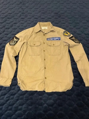 Beige skjorta från Denim & Supply - Säljer en beige overshirt från Denim & Supply med coola patchar på ärmarna och bröstet. Skjortan har två bröstfickor med knappar, klassisk krage och militärinspirerade detaljer. Tillverkad i bomull och har en avslappnad passform.