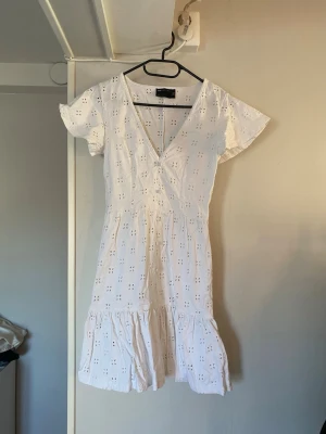 Vit omlottklänning med broderi - Söt vit kortklänning med v-ringning. Klänningen har korta ärmar, volang nedtill och är dekorerad med broderade hålmönster. Perfekt för varma dagar och sommarens alla tillfällen.