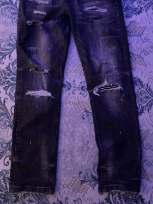 Svarta slitna skinny jeans - Säljer ett par svarta skinny jeans med flera slitningar och hål på benen. Jeansen har en tvättad look och vita färgstänk för en extra edgy stil. Perfekta för dig som gillar streetwear och vill ha en tuff vibe.