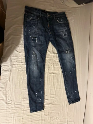 Mörkblå distressed jeans med stänk - Säljer ett par mörkblå jeans med slim fit och slitna detaljer samt vita färgstänk. Jeansen har klassisk femficksmodell, knappgylf och patchade hål på framsidan. Perfekta för en trendig och avslappnad stil.