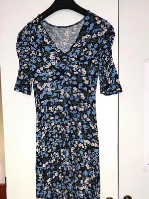 Blommig blå klänning med v-ringning - Söt vadlång klänning med blått och vitt blommönster, v-ringad hals och korta ärmar. Figurnära passform med lätt puff vid axlarna. Perfekt för vår och sommar. Material bomull 