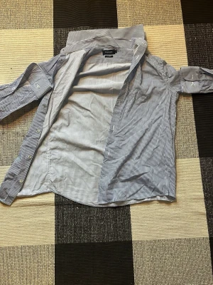 Randig skjorta från Asos - Klassisk skjorta från Asos med smala blå och vita ränder. Skjortan har lång ärm, knappar framtill och krage. Tillverkad i bomull för en bekväm känsla och snygg passform. Perfekt för en stilren och avslappnad look.