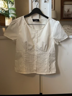 Vit blus med spetsdetaljer från Cider - Söt vit blus från Cider med korta puffärmar, knappar framtill och dekorativa spetsdetaljer längs sidorna. Blusen har rund halsringning och veckade detaljer på framsidan. Perfekt för en romantisk och trendig look. I storlek S, har använt max 4 gånger. 