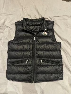Svart dunväst från Moncler - Snygg svart dunväst från Moncler med två dragkedjeförsedda fickor framtill. Västen har ett quiltat mönster och Moncler-logga på bröstet. Perfekt för lager-på-lager och håller dig varm med sin fluffiga dunstoppning. Pris kan diskuteras vid snabb affär :)