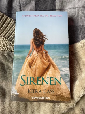 Sirenen av Kiera Cass - En ungdomsroman av Kiera Cass med ett omslag som visar en tjej i en guldig klänning vid havet. Boken handlar om Kahlen, en siren som kämpar med förbjuden kärlek och sitt öde. Perfekt för dig som gillar romantik och fantasy.