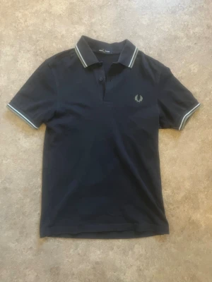 Marinblå piké från Fred Perry - Marinblå pikétröja från Fred Perry med klassisk krage och vita ränder på krage och ärmslut. Broderad logga på bröstet och knappar vid halsen. Tillverkad i mjuk bomull för en skön känsla och snygg passform.