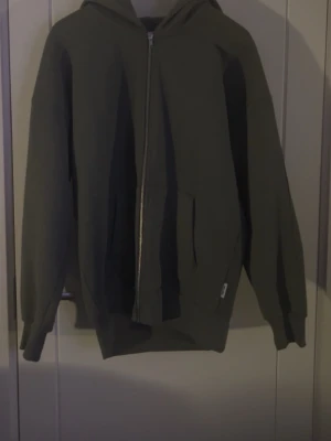 Olivgrön zip hoodie med huva - En enkel och stilren olivgrön hoodie med dragkedja framtill och stor ficka på magen. Hoodien har en klassisk huva och ribbade muddar vid ärmar och nederkant. Perfekt för en avslappnad look och passar till det mesta.