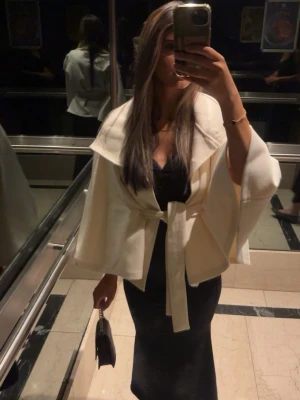 Vit poncho kappa - Stilren vit kappa med bred krage och knytskärp i midjan. Kappan har vida ärmar och en elegant, lös passform som ger en modern siluett. Perfekt att slänga över en outfit för en chic look. Materialet ser ut att vara mjukt och något tjockare tyg.