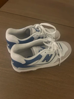 New Balance 550 vita/blå sneakers - Snygga New Balance 550 sneakers i vitt och blått med detaljer i mocka och mesh. Klassisk retrostil med gummisula och tydlig N-logga på sidan. Perfekta för dig som gillar streetwear och sportig vibe.