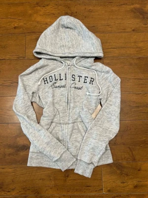 Grå zip hoodie från Hollister - Ljusgrå hoodie med dragkedja från Hollister. Broderad text 'Hollister Sunset Coast' på bröstet. Mjuk bomullsblandning, två fickor fram och justerbar huva med snören. Perfekt för en avslappnad och sportig stil.