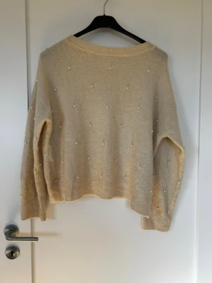 Beige stickad tröja med pärlor - Mysig beige stickad tröja med dekorativa pärlor över hela framsidan och ärmarna. Tröjan har en avslappnad passform, rund halsringning och ribbade muddar vid ärmslut och nederkant. Perfekt för dig som gillar en enkel men ändå trendig look.