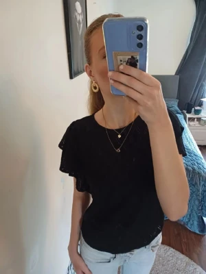 Svart topp med volangärmar - Snygg svart topp med volangärmar och diskret rutmönster i tyget. Rund halsringning och luftig passform, perfekt till jeans eller kjol. 