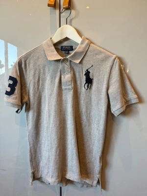 Grå pikétröja från Polo Ralph Lauren - Klassisk grå pikétröja från Polo Ralph Lauren med korta ärmar, krage och knappar framtill. Stor broderad logga på bröstet och siffra på ärmen. Tillverkad i mjuk bomull som känns skön mot huden.