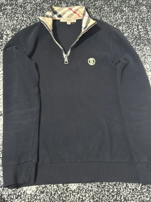 Svart Burberry halfzip med mönstrad krage (Passar till S) - Snygg svart halfzip-tröja från Burberry med klassiskt rutmönstrad krage och dragkedja. Tröjan har ribbade muddar och en diskret Burberry-logga på bröstet. Perfekt för dig som gillar stilrena plagg med exklusiva detaljer. 