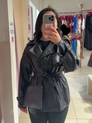 Svart trenchcoat i skinnimitation - Snygg svart trenchcoat i skinnimitation med bälte i midjan och klassisk dubbelknäppning. Jackan har breda slag, axelklaffar och justerbara spännen vid ärmsluten. Perfekt för dig som vill ha en edgy och trendig look.