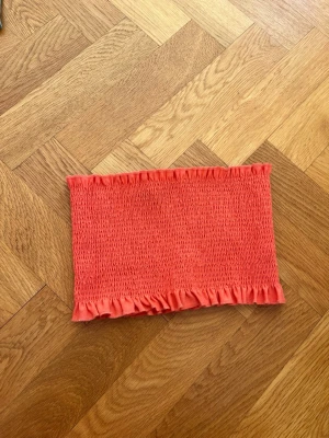 Korallfärgad bandeau tubtopp - Säljer en snygg korallfärgad bandeau tubtopp med smockad struktur och volangkant upptill och nedtill. Perfekt för varma dagar och enkel att styla med jeans eller kjol.