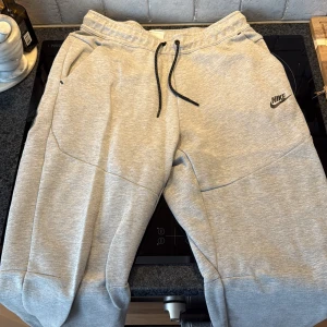 Grå mjukisbyxor från Nike, storlek M - Snygga ljusgrå mjukisbyxor från Nike med svart logga på låret. Byxorna har dragsko i midjan, ribbade muddar vid bensluten och är tillverkade i mjuk bomullsblandning. Perfekta för chill eller träning. Klassisk och clean look. Dom är i storlek medium för män och är  köpta ifrån jdsports i Gallerian.