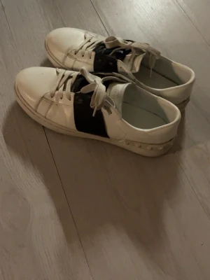Vita sneakers från Valentino - Snygga vita sneakers från Valentino med en bred svart läderdetalj över snörningen. Klassisk låg modell med vita skosnören och diskret logga på plösen. Skorna har en stilren design och passar perfekt till streetwear och casual outfits.