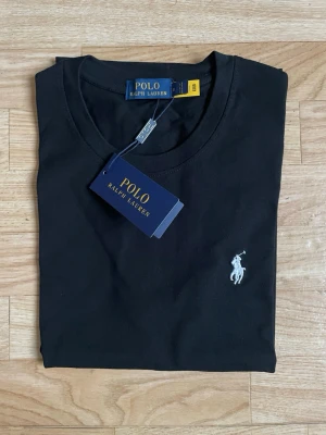 Svart t-shirt från Polo Ralph Lauren - Snygg svart t-shirt från Polo Ralph Lauren med klassisk rund hals och korta ärmar. T-shirten har den ikoniska vita polospelaren broderad på bröstet och är tillverkad i mjuk bomull för en skön känsla. Perfekt till jeans eller shorts.
