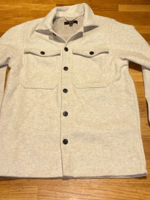 Ljusbeige overshirt från Smog - Stilren ljusbeige overshirt från Smog med klassisk krage och svarta knappar. Två stora bröstfickor med lock och knappdetaljer. Tillverkad i mjukt, filtliknande material som ger en avslappnad men ändå snygg look. Perfekt lager på lager-plagg.