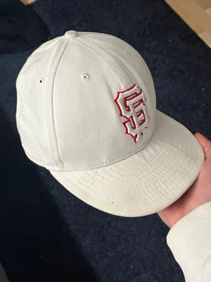 Vit New Era keps med röd logga - Snygg vit keps från New Era med broderad röd San Francisco Giants-logga framtill och MLB-logga bak. Klassisk böjd skärm och sex paneler med ventilationshål. Tillverkad i bomullstyg för en bekväm känsla.