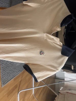 Vit Moncler piké med marinblå detaljer - Säljer en stilren vit pikétröja från Moncler med marinblå krage och ärmslut. Slim fit-modell med knappslå och klassisk Moncler-logga på bröstet. Perfekt för dig som gillar exklusiva och tidlösa plagg.