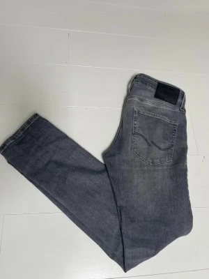 Grå jeans från Jack & Jones - Säljer ett par grå jeans från Jack & Jones med klassisk femficksmodell och svarta nitar. Jeansen har en rak passform och är tillverkade i ett mjukt denimtyg med lätt tvättad look. Perfekta för en avslappnad och stilren outfit. Storlek 28 - 30